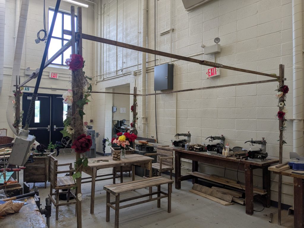 Outdoor living display with an A-frame shelter, table, benches, planter boxes, and floral decorations built from spacer-stick wood.