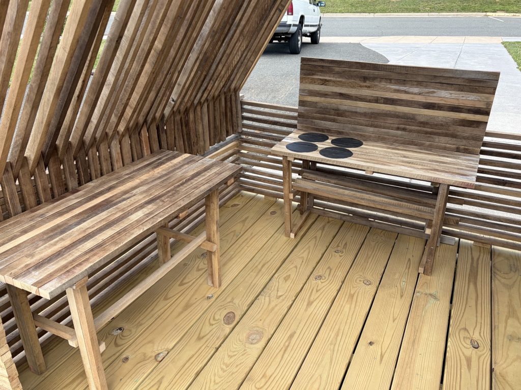 Interior of an A-frame outdoor sitting structure with a wood deck, slatted walls, bench, small table, and round coasters.