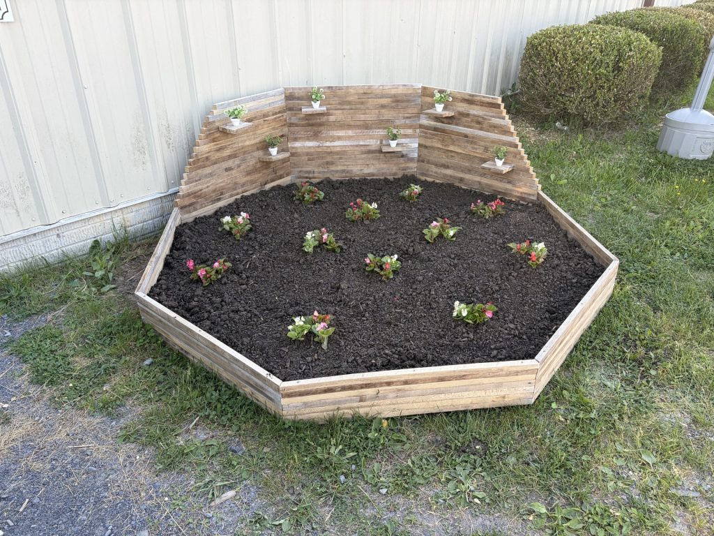 Hexagonal raised flower bed with horizontal spacer-stick slats, angled privacy panels, plant shelves, soil, and flowers.