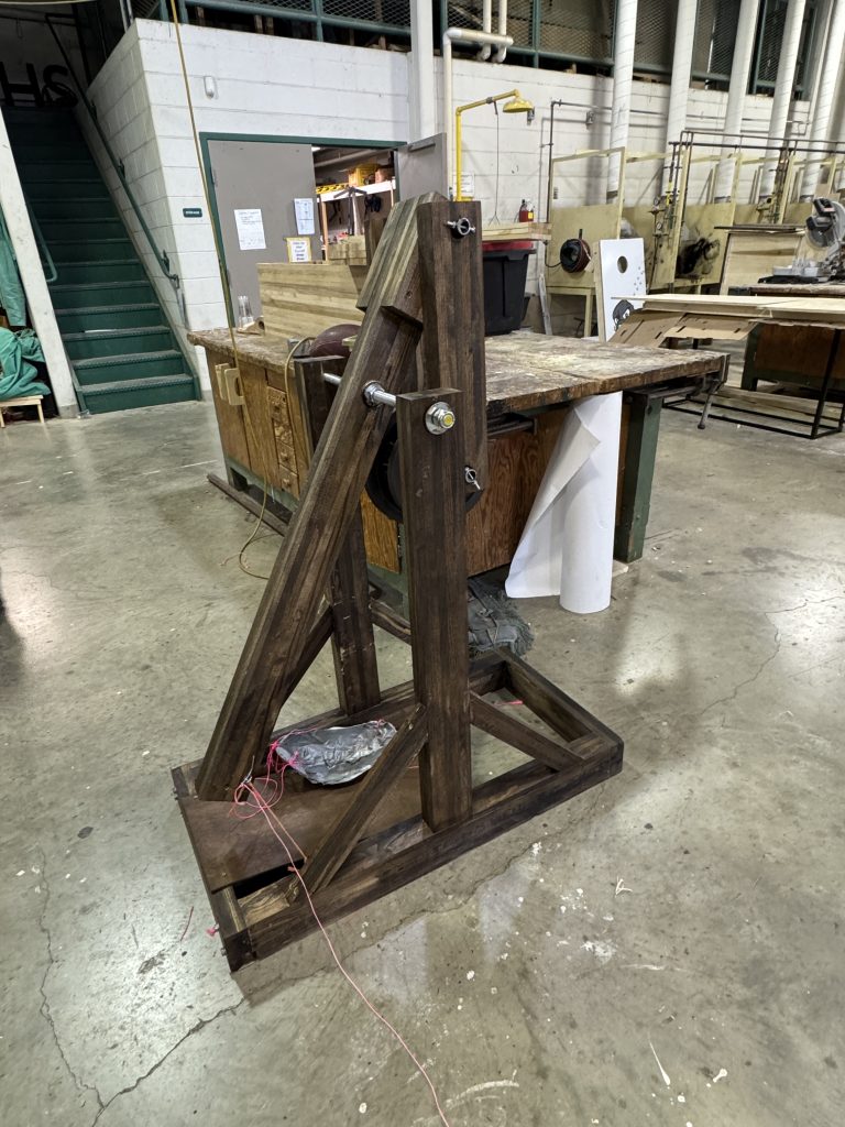 Large dark-stained wooden trebuchet with a triangular base, upright frame, throwing arm, and hanging counterweight.