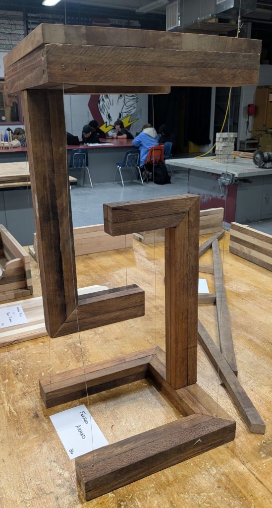 Suspended wooden sculpture with dark spacer-stick blocks and geometric pieces hanging from clear lines inside a rectangular frame.