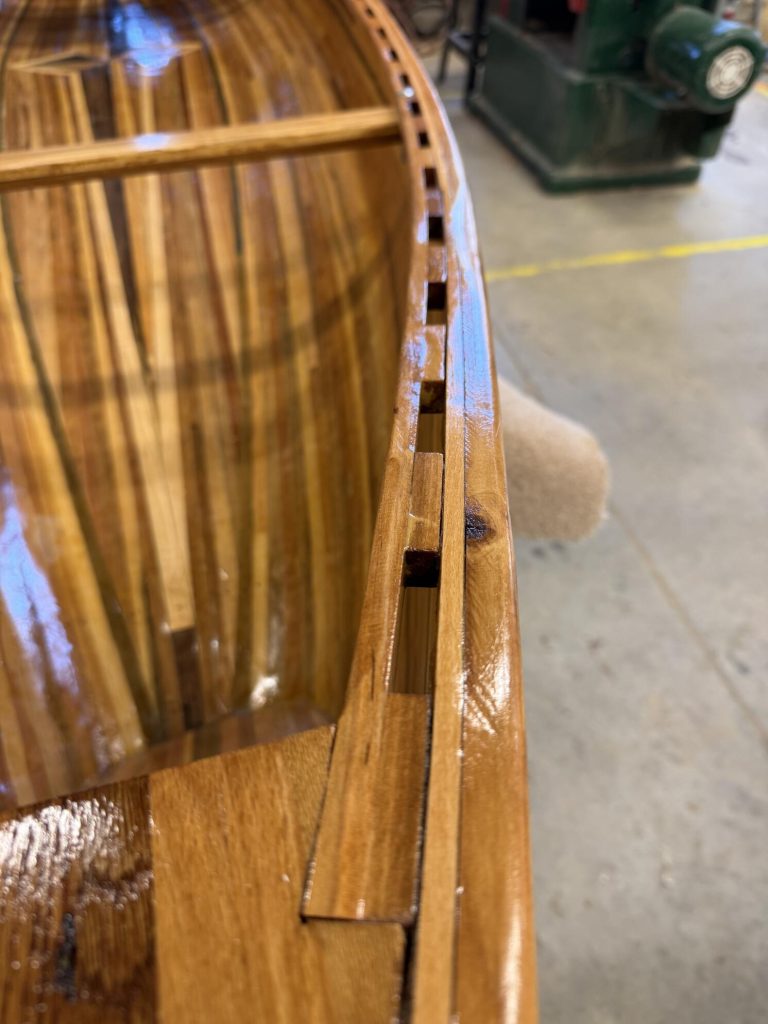 Close-up of a glossy wooden canoe showing the shaped gunwale, spacer-stick strip construction, and side joinery.
