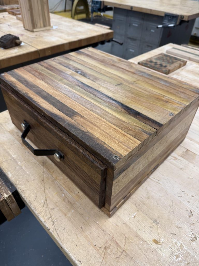 Close-up of a striped wooden storage box or portable case with a framed lid, metal handle, and mixed-tone spacer-stick panels.